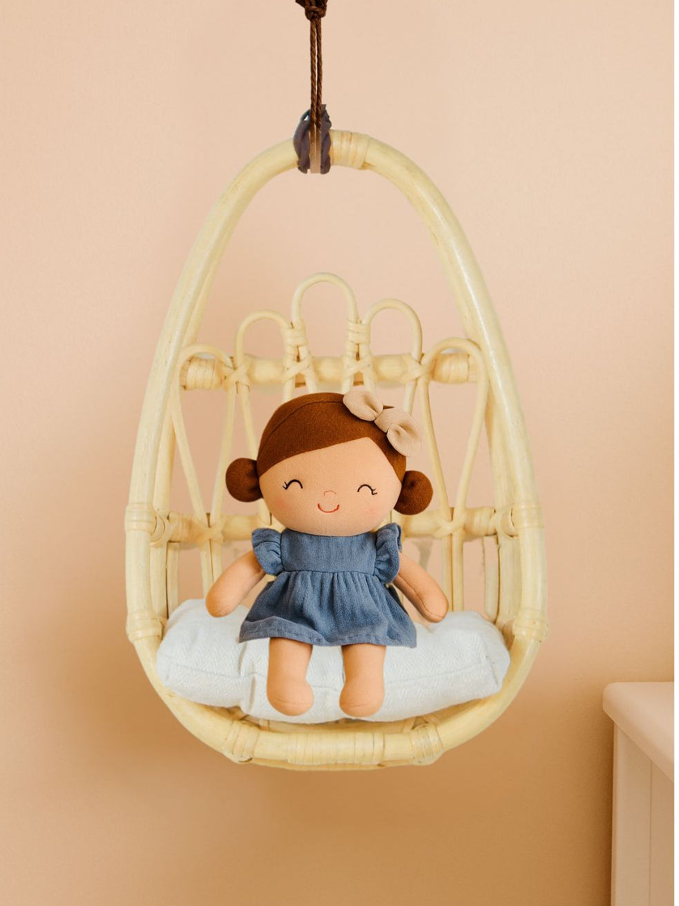 Plush doll in a wicker chair against a beige wall. Handcrafted rattan wall chair for dolls, bringing a natural boho touch to nursery and playroom decor.