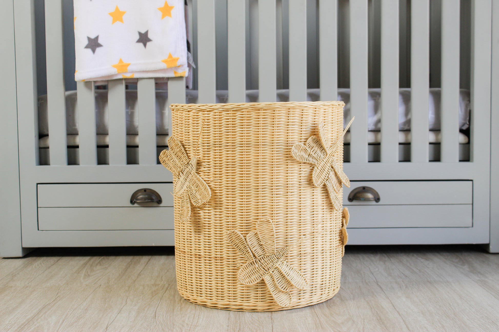 Wicker laundry basket sits in front of a gray baby crib. Insect, butterfly and dragonfly theme hamper, toy storage basket. 