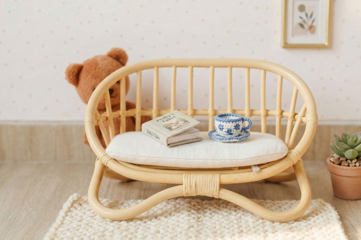 A detailed, eye-level photograph of a miniature, handcrafted rattan doll couch with a white linen cushion. It is set in a peaceful nursery on a woven rug. In the background, a brown plush teddy bear is visible, alongside a small potted succulent and framed wall art, illustrating sophisticated nursery decor and gifts for kids.
