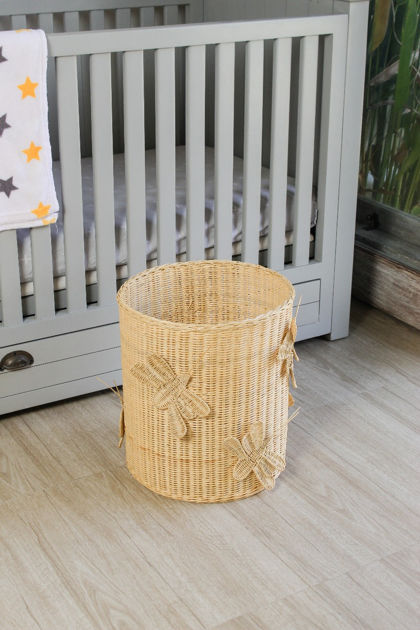 Wicker laundry basket with dragonfly design on a wooden floor next to a crib. Rattan made toy storage for kids toys. Basket for toys