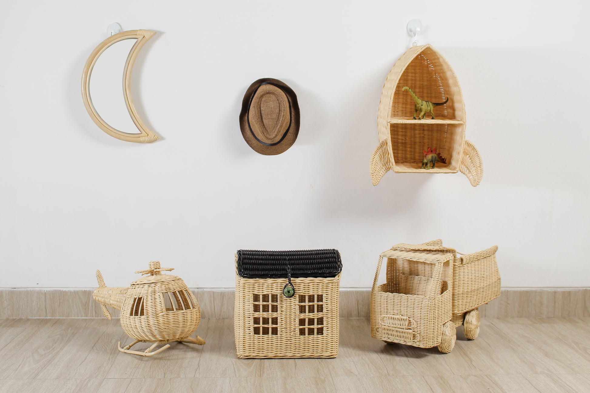 A hand-made moon shaped wall mirror sits on a wall next to a hat and a rocket ship shaped shelf. On the floor sits a toy helicopter, a basket house and a toy truck. 