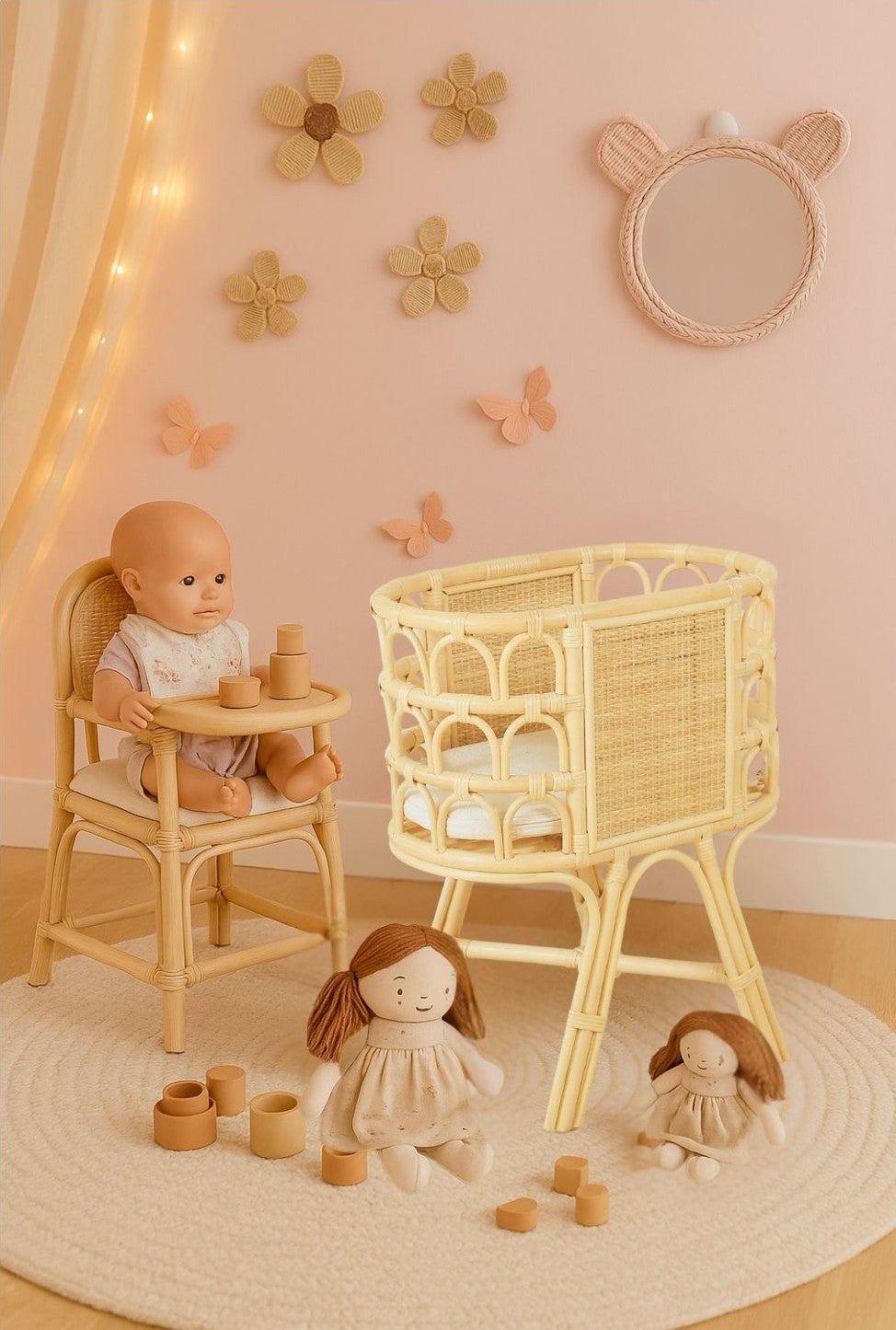 Children's room with wicker high chair, crib, and toys on a light pink wall with decorative elements. A doll's crib sits in front of two dolls. Flower wall decor appears on the wall. 