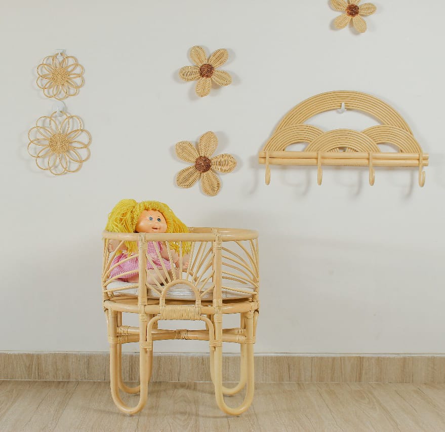 A cabbage patch doll sits in a hand-made dolls bed. On the wall is rattan wall decor flowers with a rainbow hanging rack. It is cute and pretty