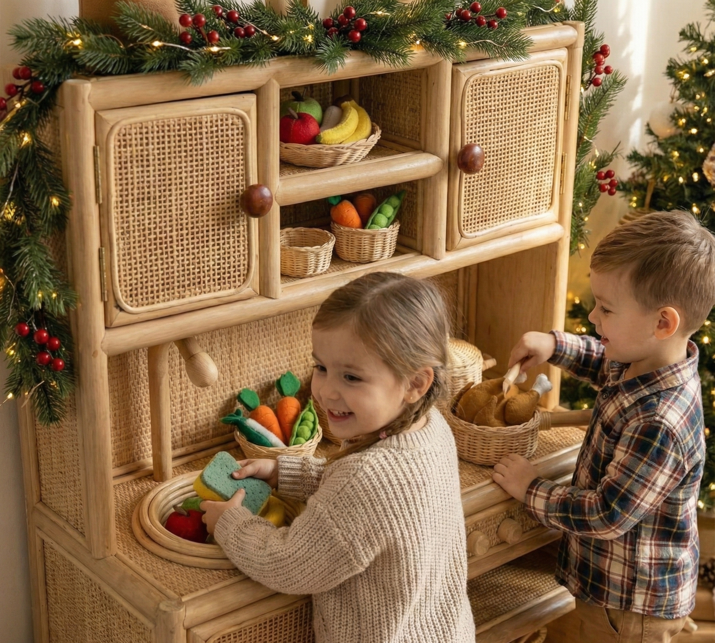Two children playing with toy vegetables in a wicker play kitchen set against a Christmas tree. Best toddler Christmas present 2025. 