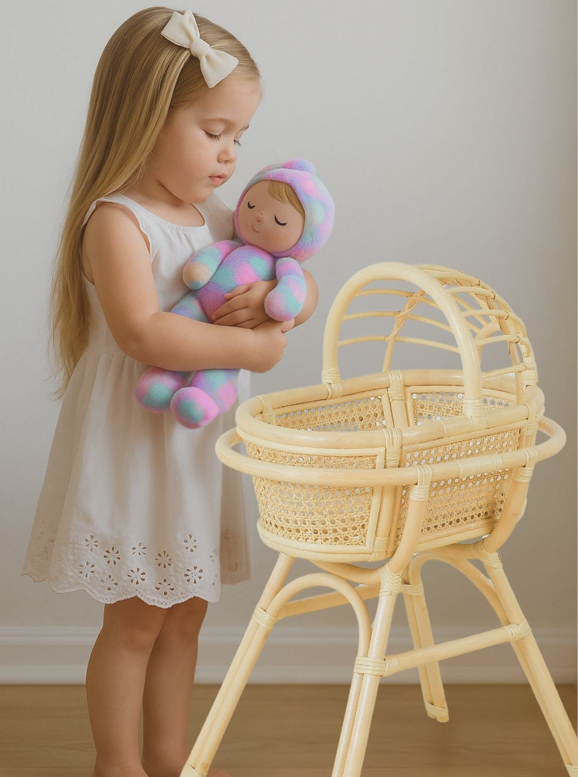 Young girl holding a colorful baby doll next to a wicker bassinet. Doll bed