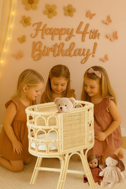 A teddy bear sits in a rattan made dolls bassinet or a dolls crib or a doll bed. Three girls dressed in pretty dresses look at it with two dolls also sitting on the ground. 