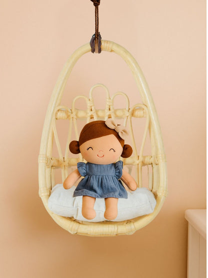 Plush doll in a wicker chair against a beige wall. Handcrafted rattan wall chair for dolls, bringing a natural boho touch to nursery and playroom decor.