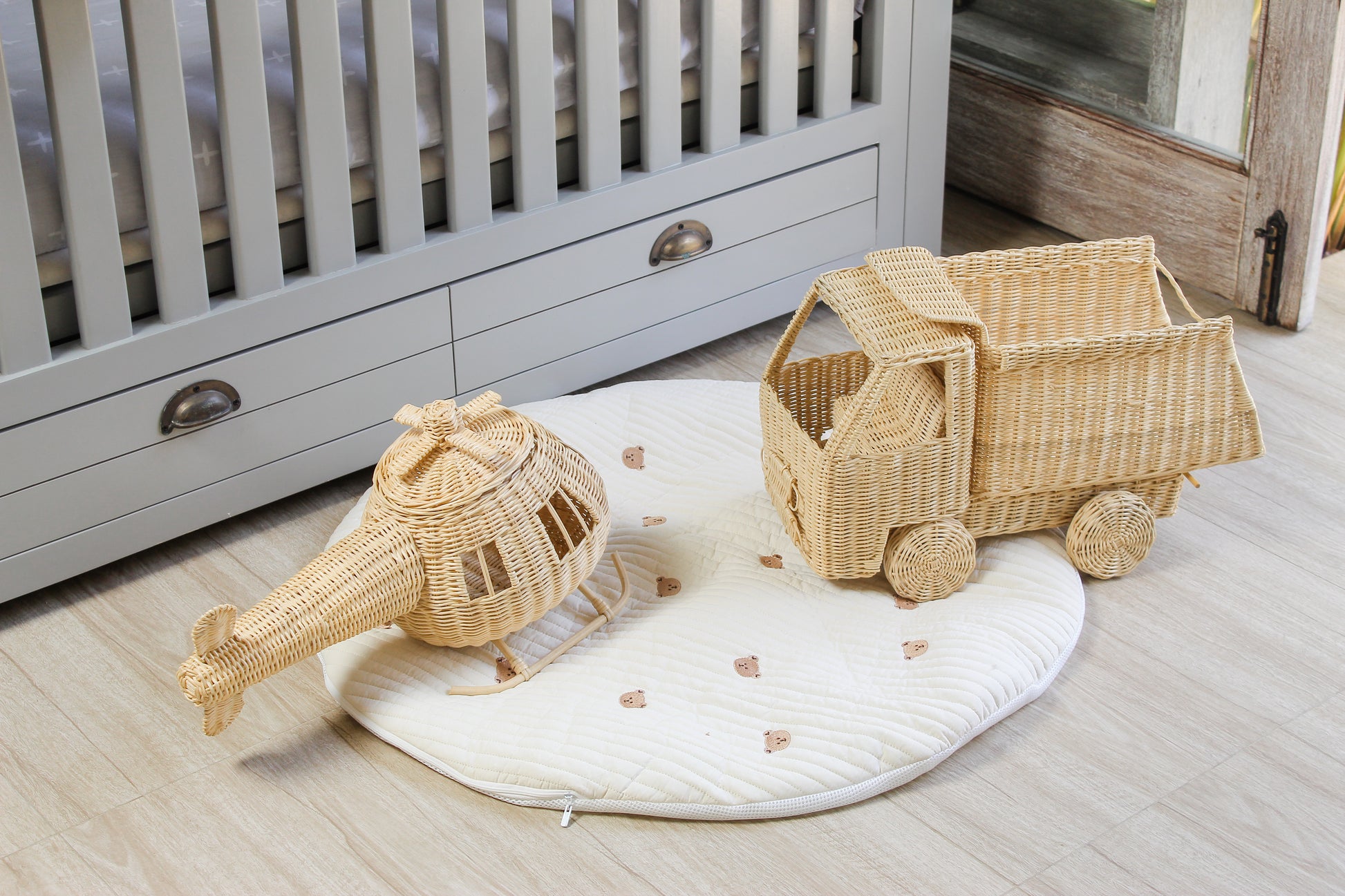 Wicker toy truck and helicopter on a white mat in a nursery with a crib in the background.
