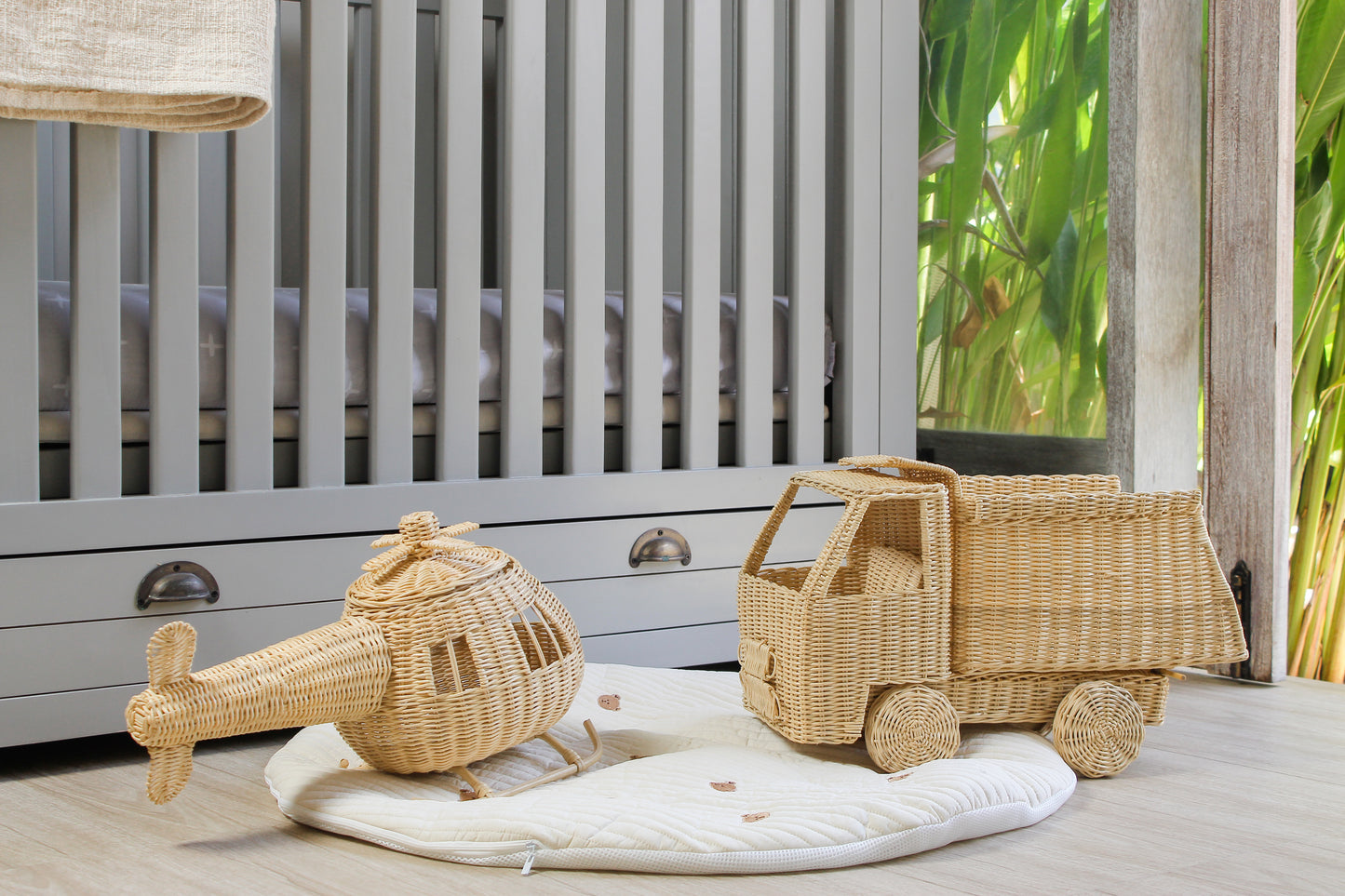 Wicker toy truck and helicopter on a white mat in front of a crib with a natural outdoor background.