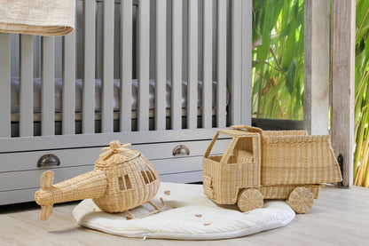 Wicker toy truck and helicopter on a white mat in front of a crib with a natural outdoor background.